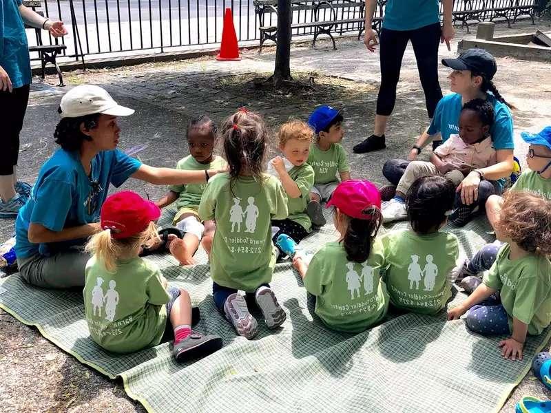 group of toddlers on a blanket in a park, with a couple of Barnard students